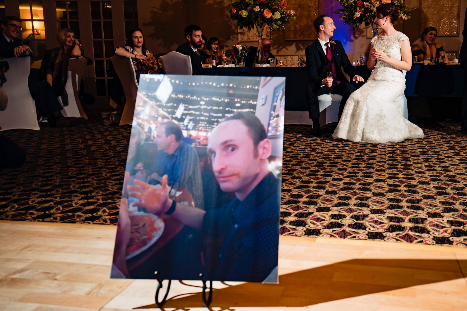 A colorful picture, focused on a printed picture of a man covered in buffalo wing sauce with a bride and groom laughing hysterically behind the picture during a winter wedding reception at The Elms. 