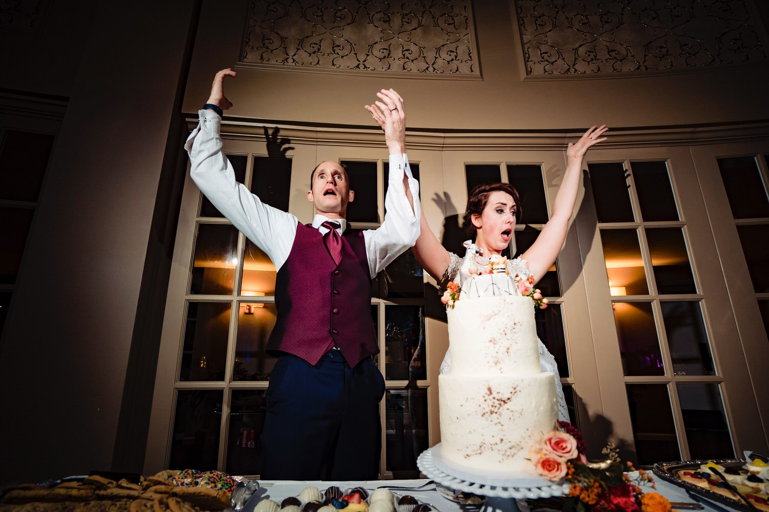 A candid picture of a bride and groom throwing their arms up and yelling in celebration just after they cut their wedding cake during their wedding reception at The Elms. 