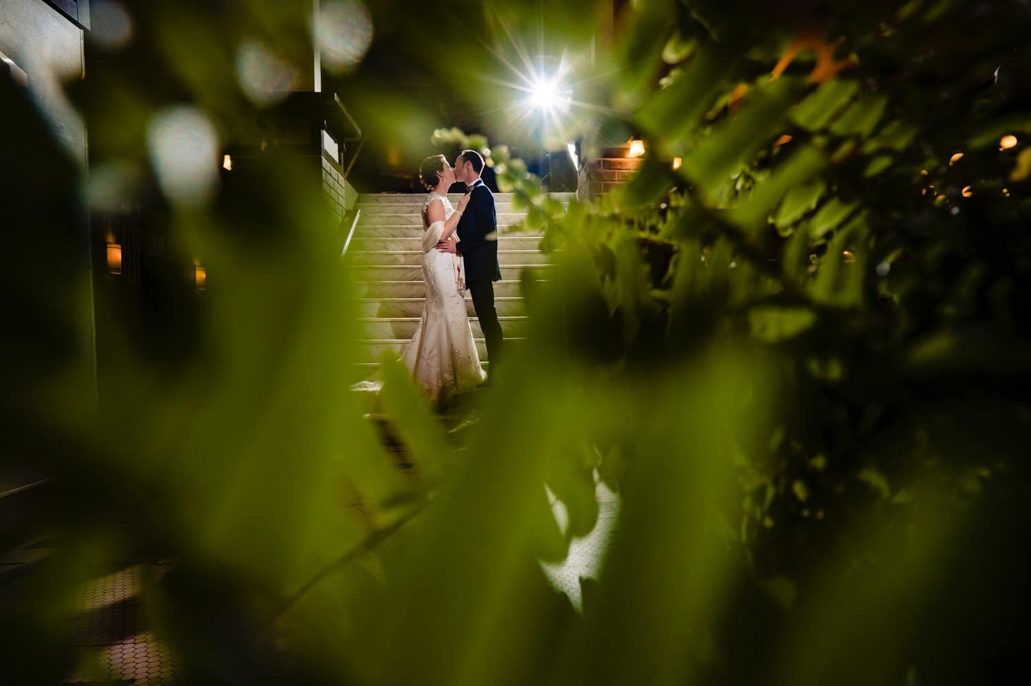 A colorful portrait taken through the leaves of a green plant of a bride and groom standing on a set of stairs in the lobby of The Elms Hotel, sharing a kiss at the end of their wedding day. 