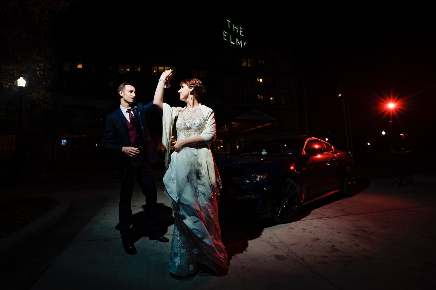 A candid picture of a bride and groom dancing in front of a dark blue mustang parked in front of The Elms Hotel on a winter wedding day. 