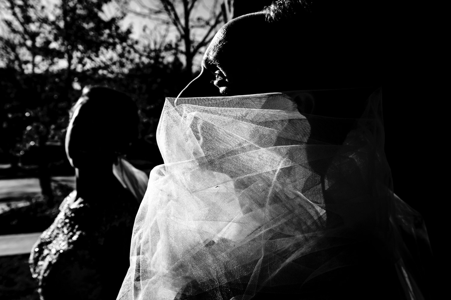 A dramatic black and white candid picture of a bride standing with her hands on her hips as her groom attempts to wrangle her veil on their wedding day. 