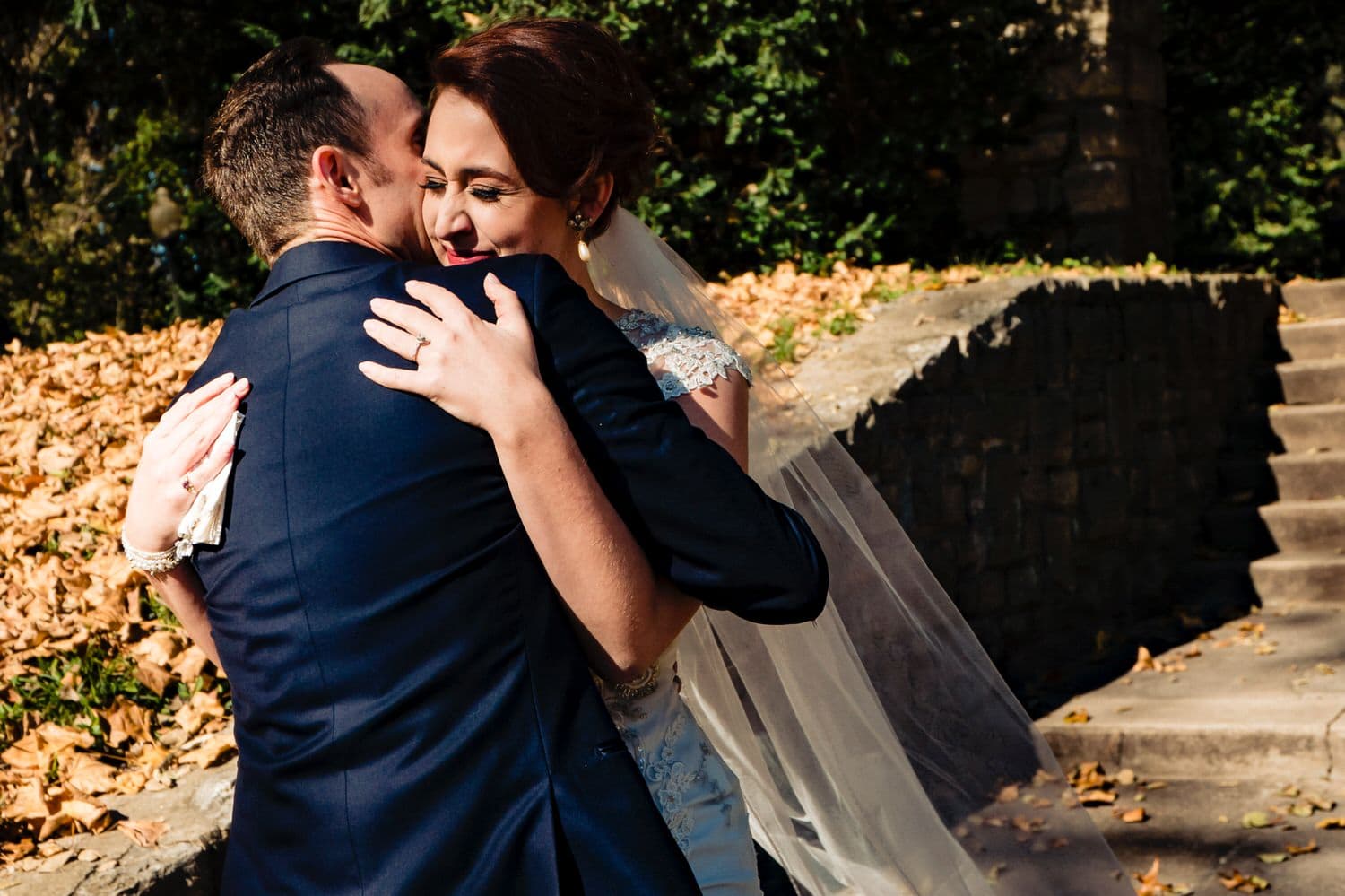 A colorful, candid picture of a bride hugging a groom in a blue suit as he kisses her on the cheek during their first look on their wedding day at The Elms. 