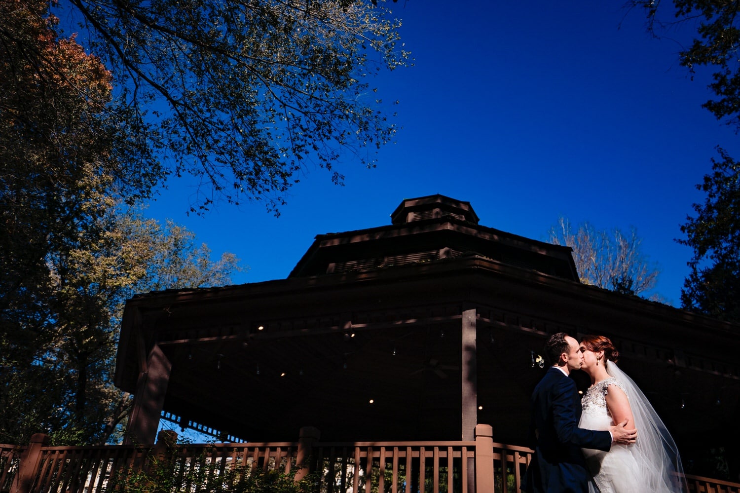 A colorful, candid picture of a bride and groom sharing a kiss as they stand in front of the gazebo at The Elms on their wedding day. 
