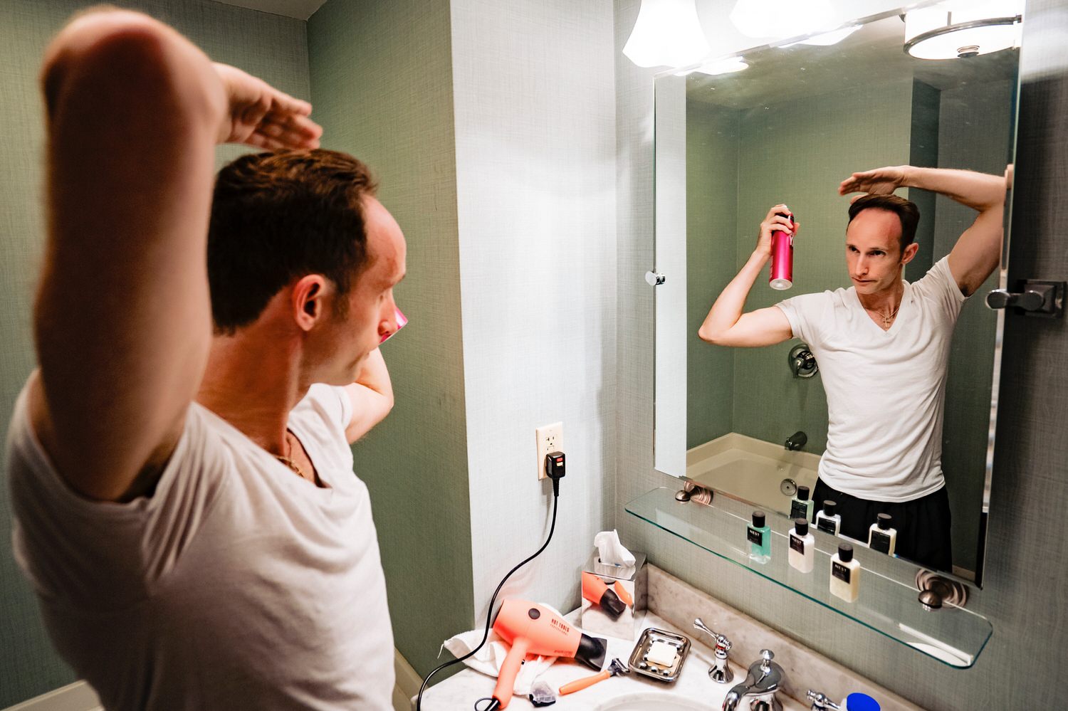 A colorful, candid picture of a groom spraying hair spray on his hair in a bathroom on his wedding morning at The Elms. 