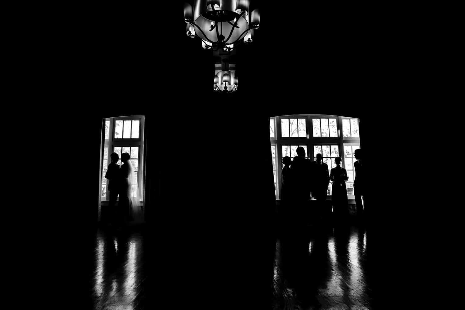 A dramatic black and white portrait of a bride and groom silhouetted in one window of a ballroom at The Elms, with their bridal party silhouetted in the other window. 
