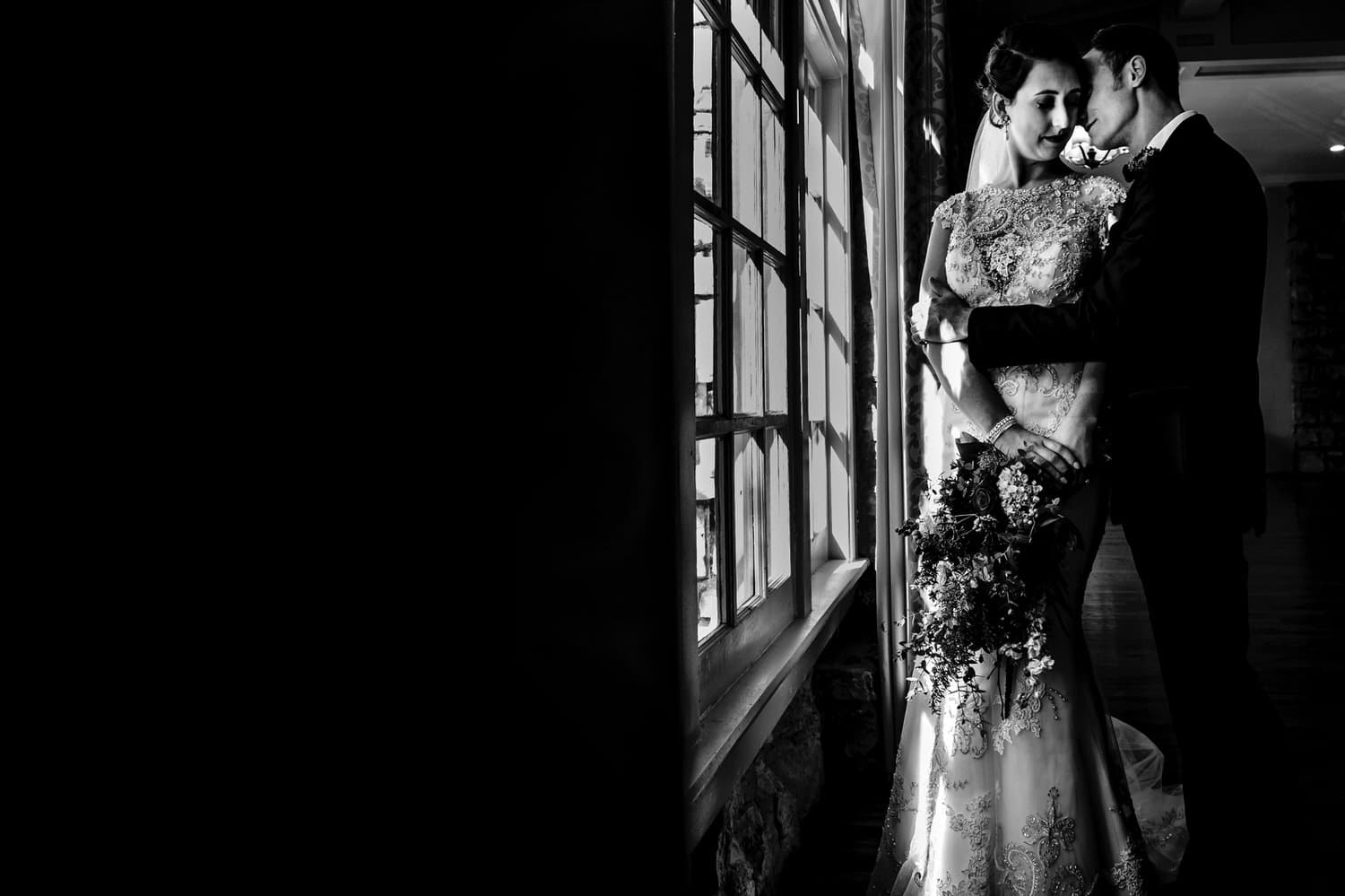 A bold black and white portrait of a groom leaning in to kiss his bride's cheek as they stand by a window on their winter wedding day at The Elms. 