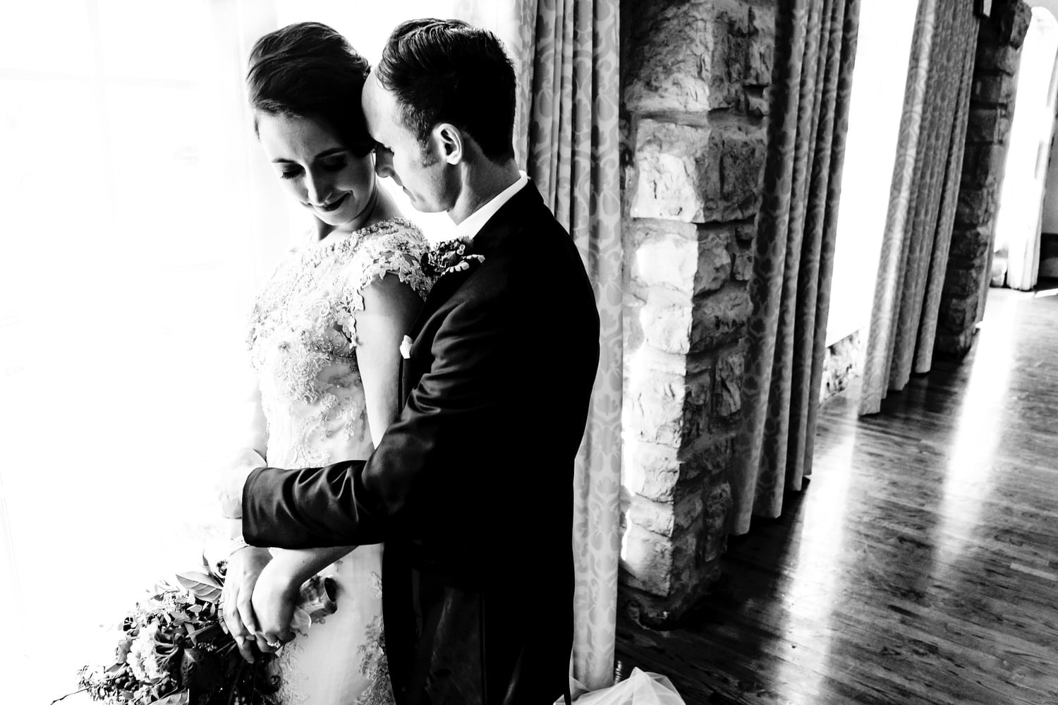 A bold black and white portrait of a groom leaning in to kiss his bride's cheek as they stand by a window on their winter wedding day at The Elms. 