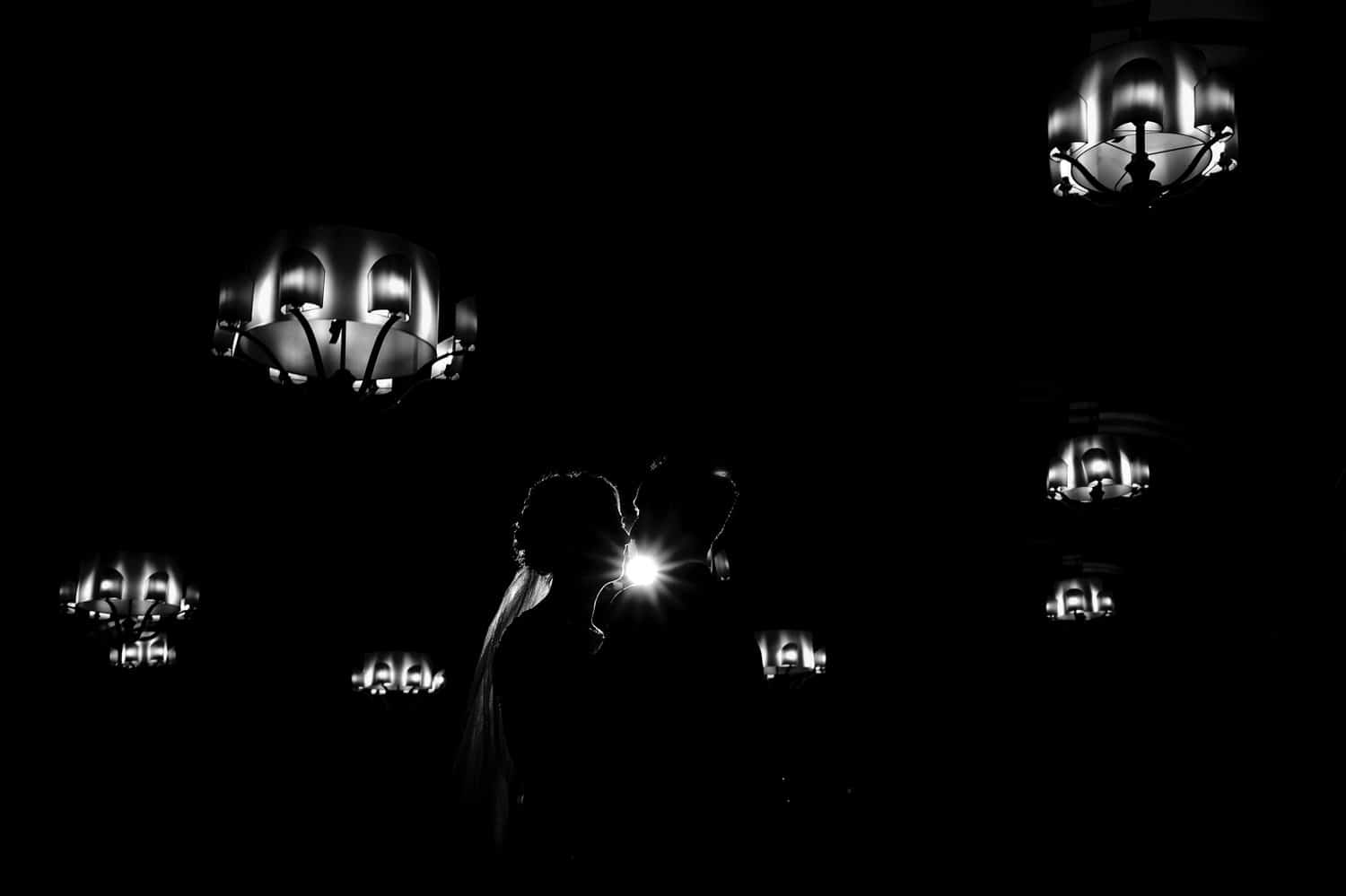 A dramatic black and white silhouetted portrait of a man and woman leaning in to share a kiss, the chandeliers hanging in the ceiling visible above them on their winter wedding day at The Elms. 