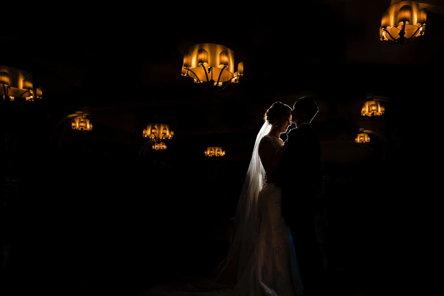 A dramatic color silhouetted portrait of a man and woman leaning in to share a kiss, the chandeliers hanging in the ceiling visible above them on their winter wedding day at The Elms. 