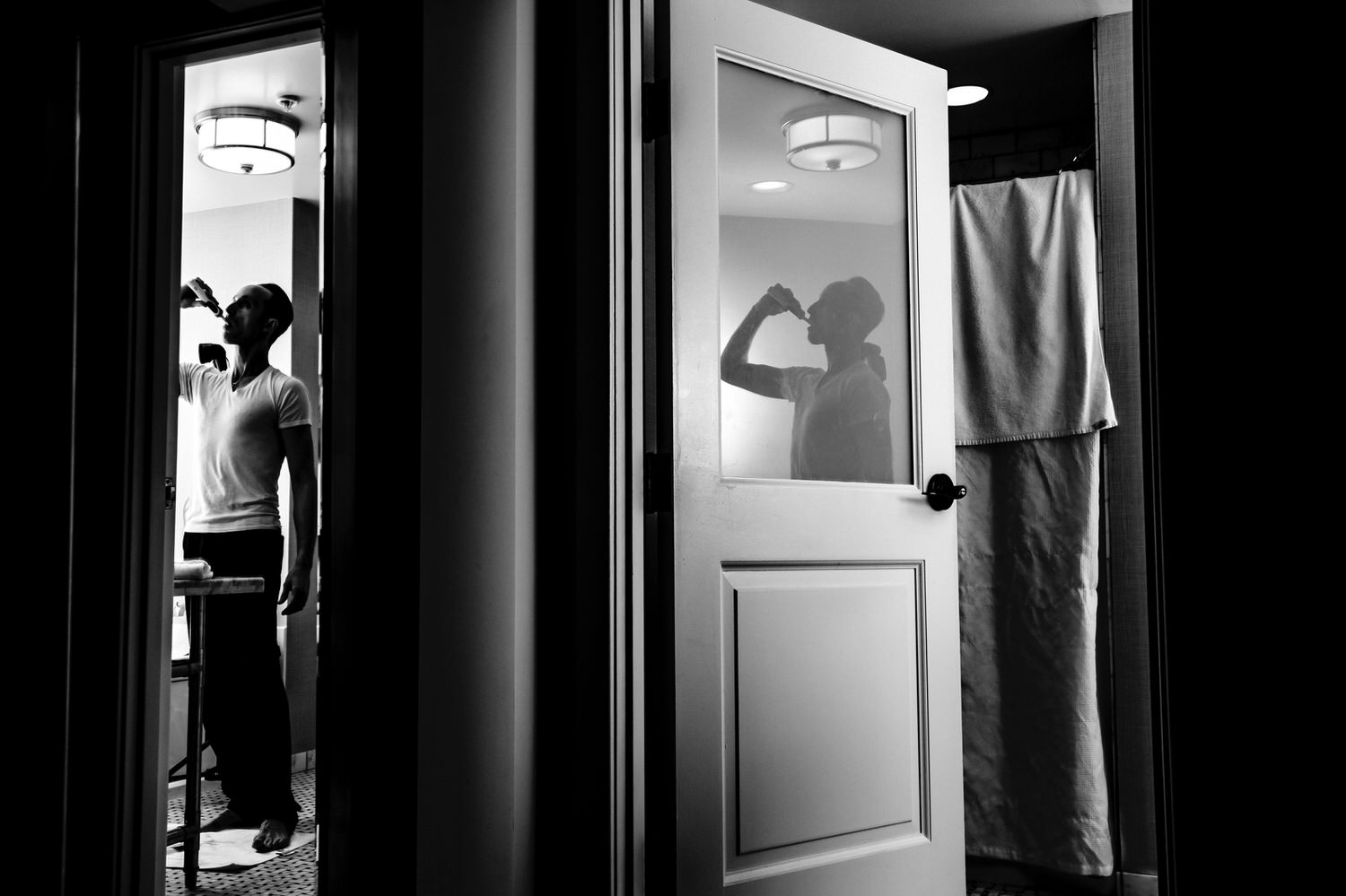 A candid black and white picture of a man brushing his teeth, his reflection visible in a door and mirror of a hotel room at The Elms. 