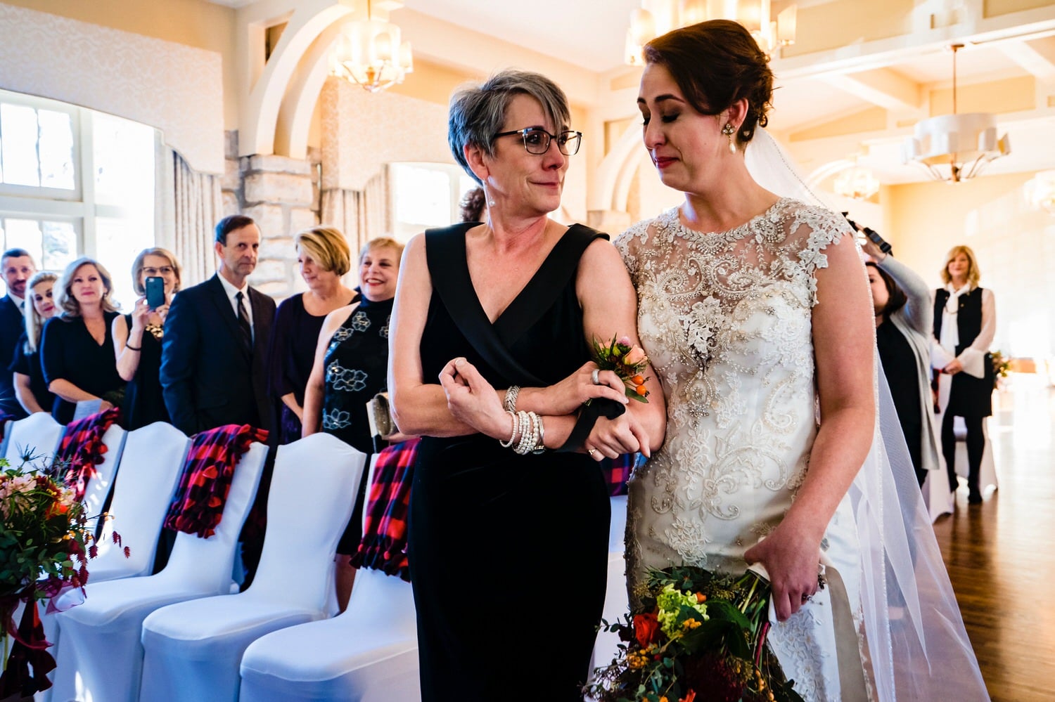 A colorful, candid picture of a bride's mom trying to comfort the bride as happy tears roll down both of their faces as they walk down the aisle for a winter wedding ceremony at The Elms. 