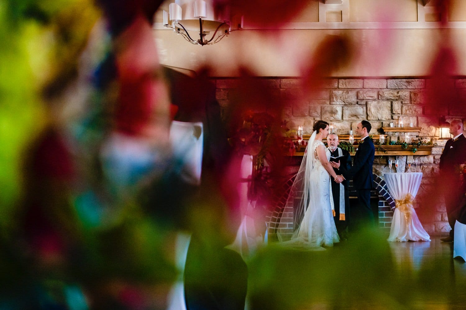 A picture of a bride and groom holding hands, exchanging vows, taken through a red floral arrangement during a winter wedding ceremony at The Elms. 