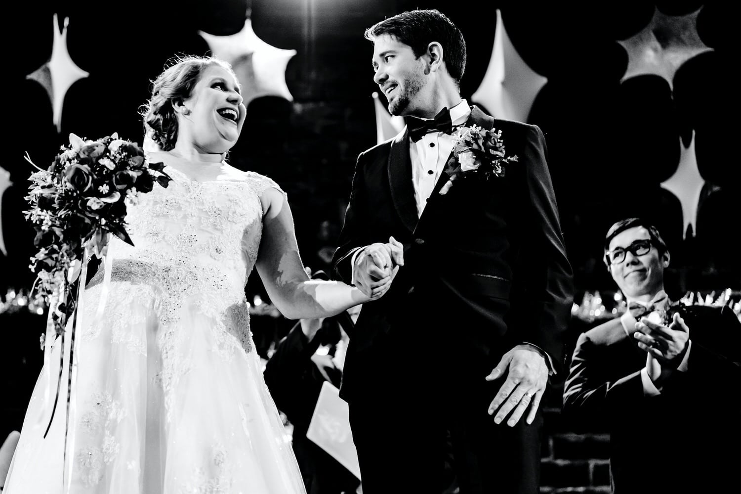A candid black and white picture of a bride holding a bouquet and a groom's hands as the couple smiles at each other in front of a backdrop of white snowflakes hanging from the ceiling of the Vox theater at the end of a wedding ceremony near Christmas time in Kansas City. 