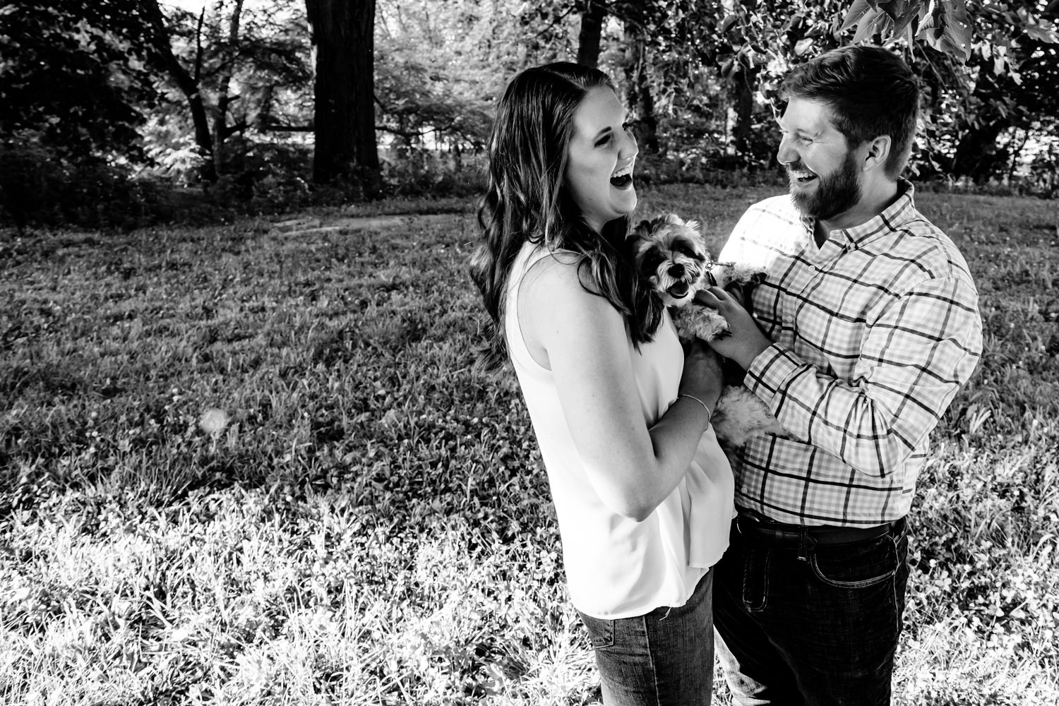 A candid black and white picture of an engaged couple laughing and holding their small dog during a Kansas City engagement session. 
