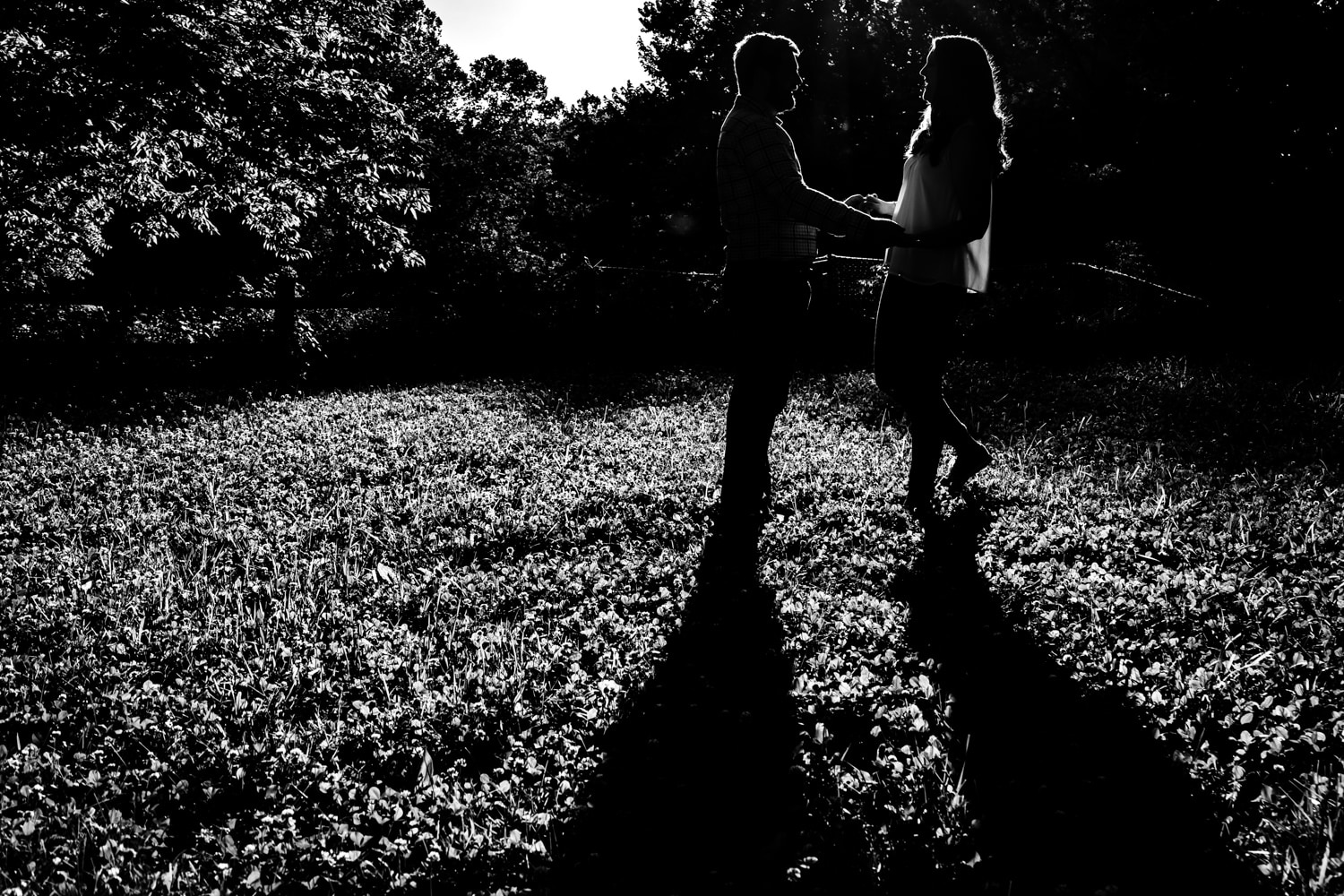 A black and white, semi-silhouetted portrait of an engaged couple holding hands and walking towards each other. 
