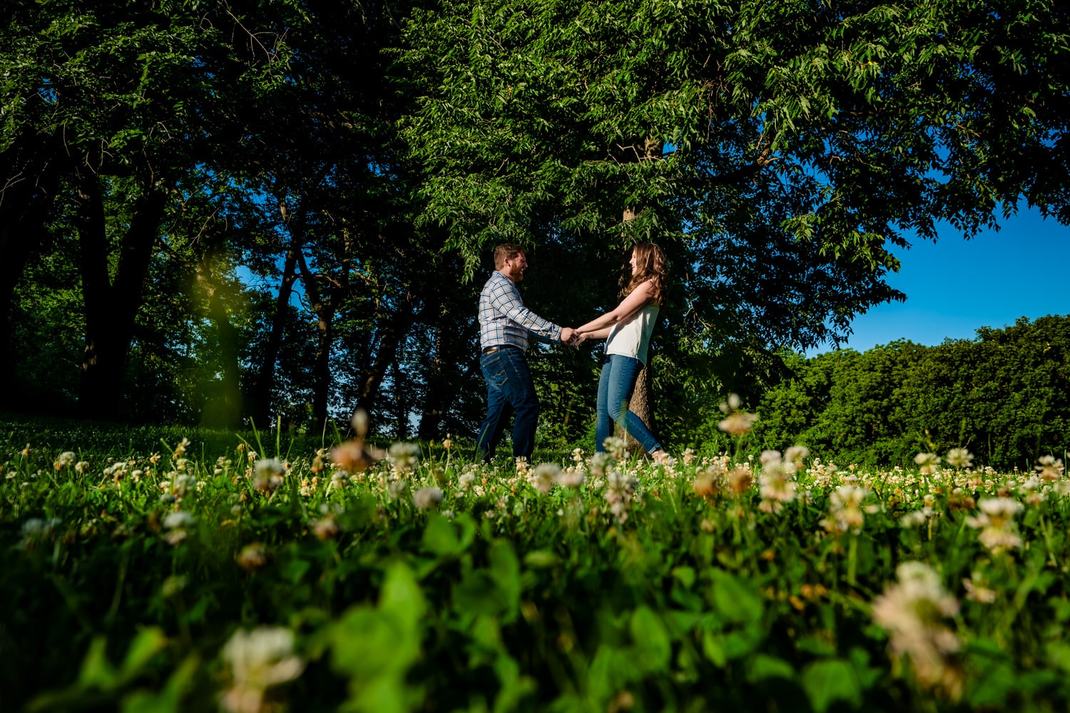 A colorful, candid picture of an engaged couple holding hands, dancing in the sun during their engagement session at Kansas City's Red Bridge Park. 
