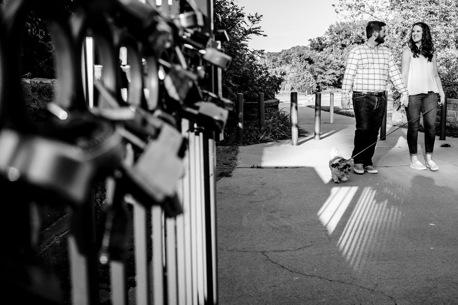 A candid black and white picture of an engaged couple walking their small dog over KC's Love Lock Bridge. 