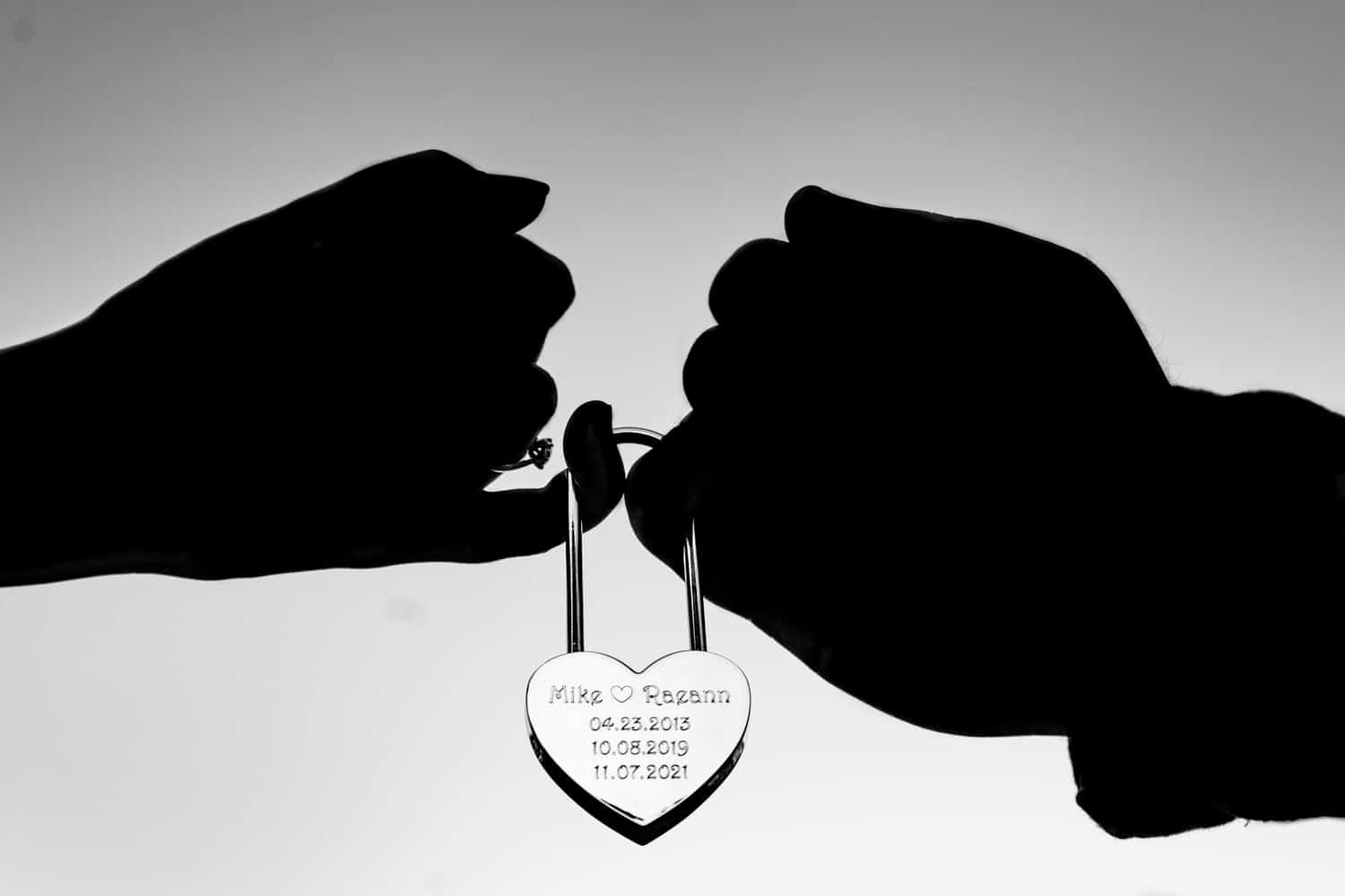 A semi-silhouetted picture of a couple's hands holding a padlock with their names and important dates on it. 