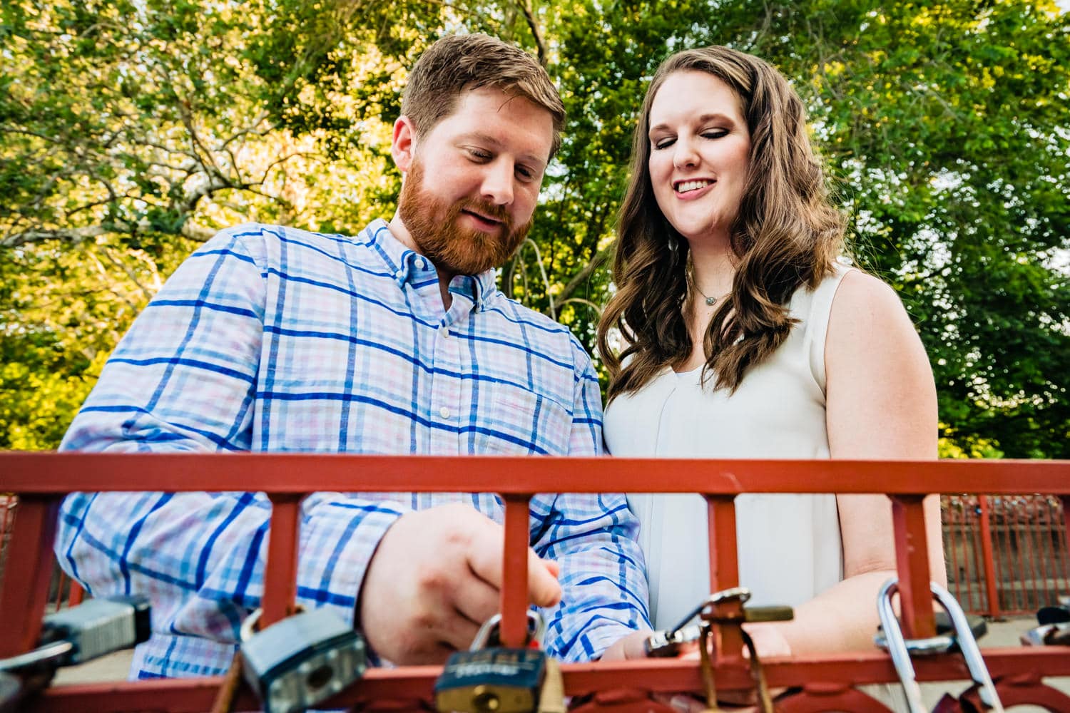 A colorful, candid picture of an engaged couple putting a padlock onto a red bridge with other padlocks during their Kansas City engagement session. 