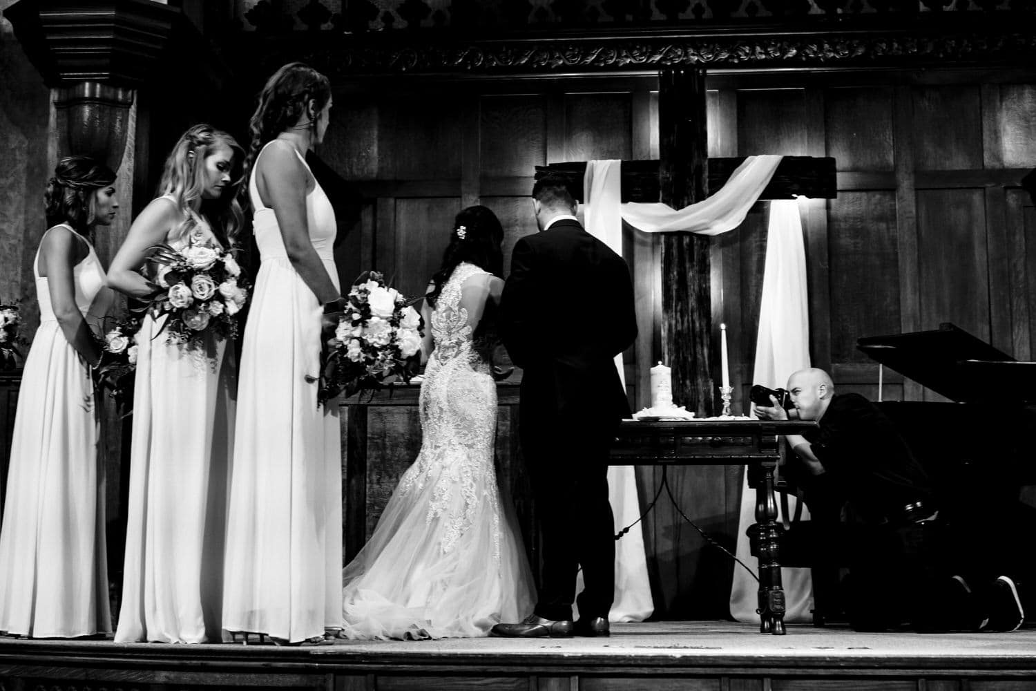 A black and white picture of a photographer leaning in to take a picture of a bride and groom lighting a unity candle during their wedding ceremony. 