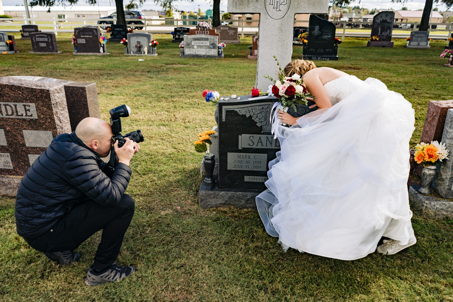 A picture of a photographer standing back and taking a photo of a bride leaning over to kiss the top of her father's grave on her wedding day. 
