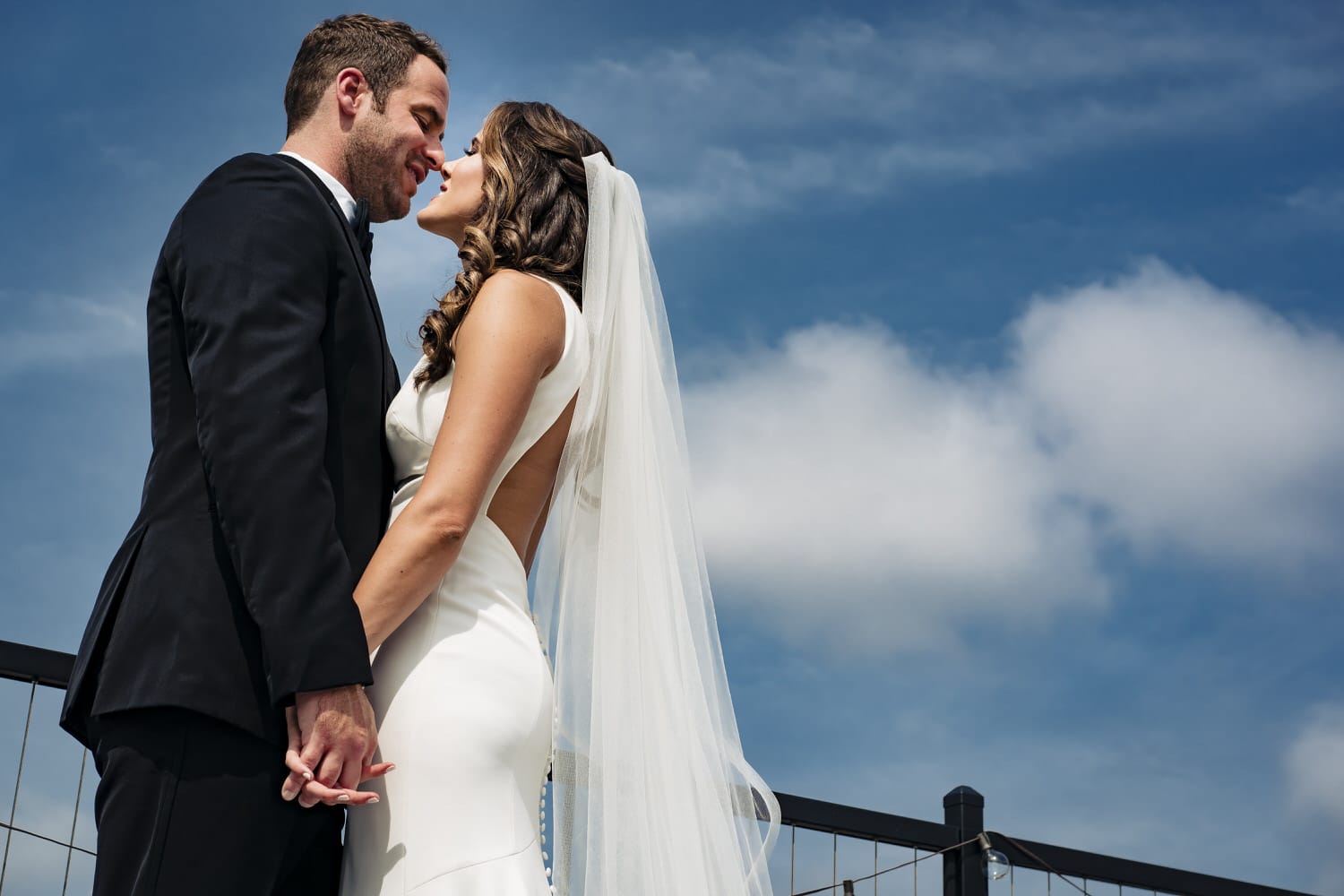 A sunny picture of a bride and groom holding hands, leaning in to share a kiss against a bright blue sky with puffy white clouds on their Kansas City wedding day. 
