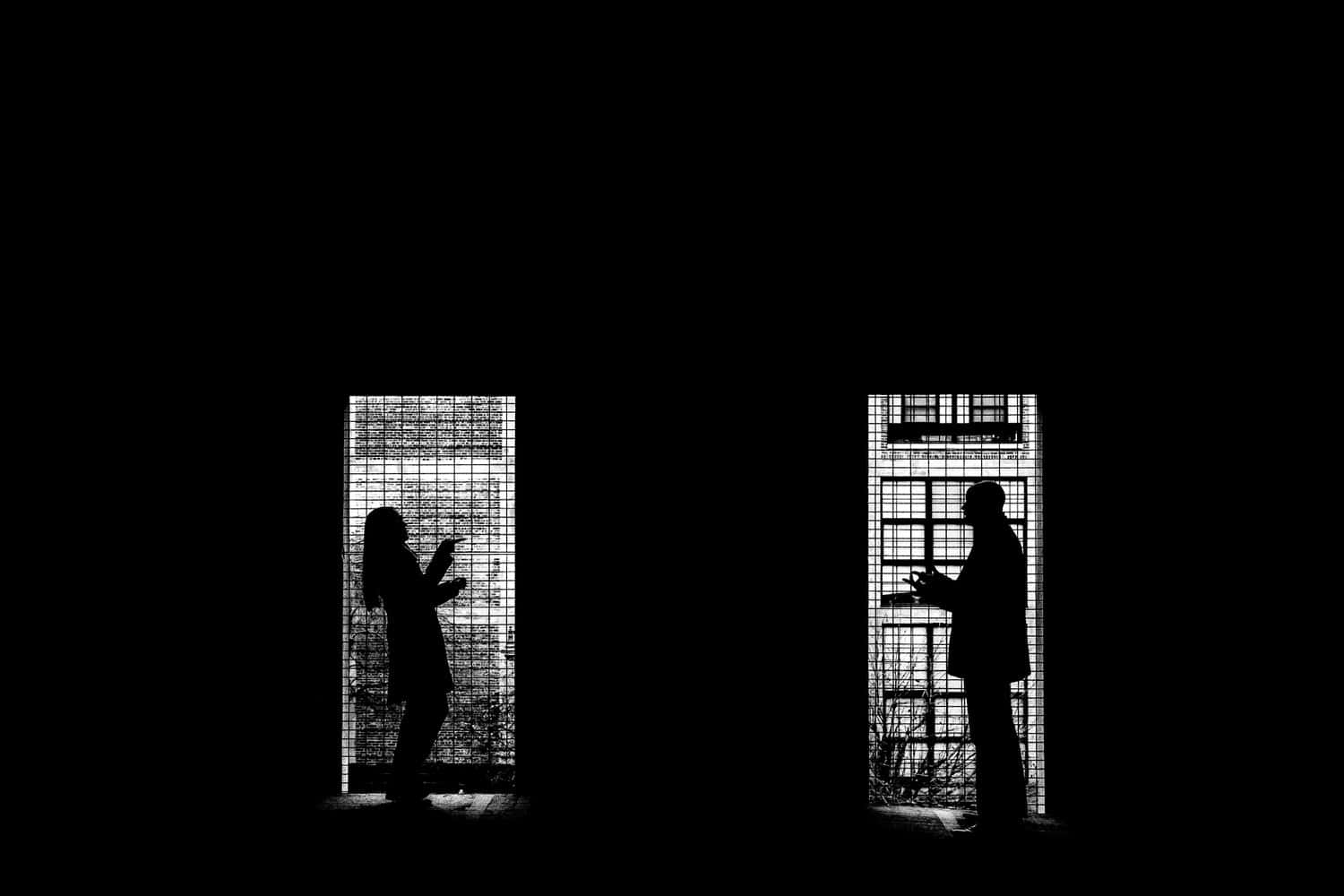 A dramatic black and white picture of two people, silhouetted in windows in an otherwise black screen making hand signals at each other and laughing during their winter downtown KC Engagement Session. 