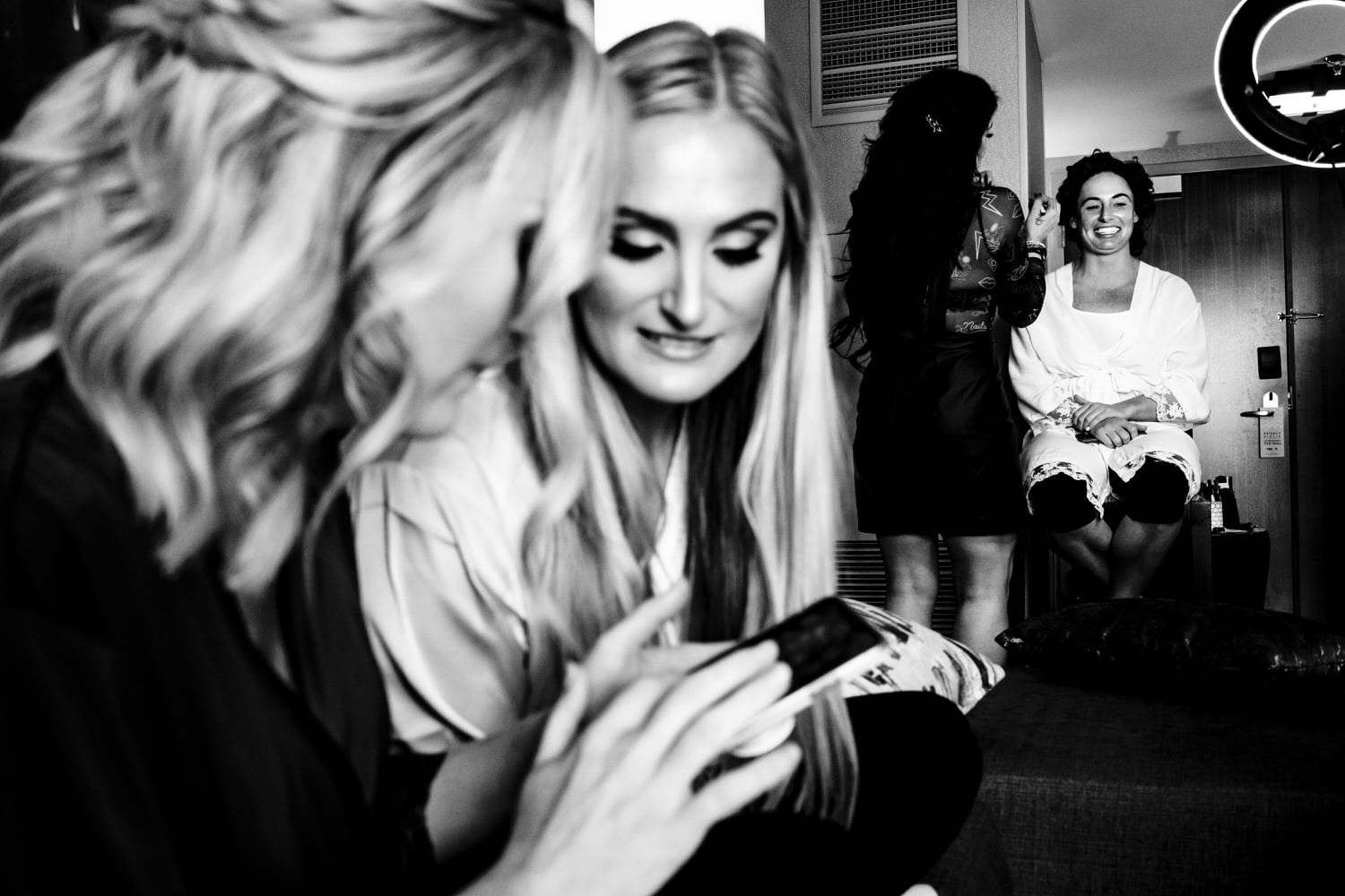 A black and white picture of a bride getting her makeup done while her sisters laugh at something on a phone. 