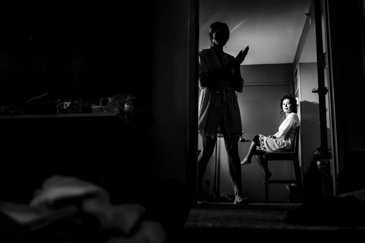 A black and white, picture of a bride sitting in a chair getting her hair and makeup done as a silhouetted bridesmaid walks by on the morning of a winter wedding. 