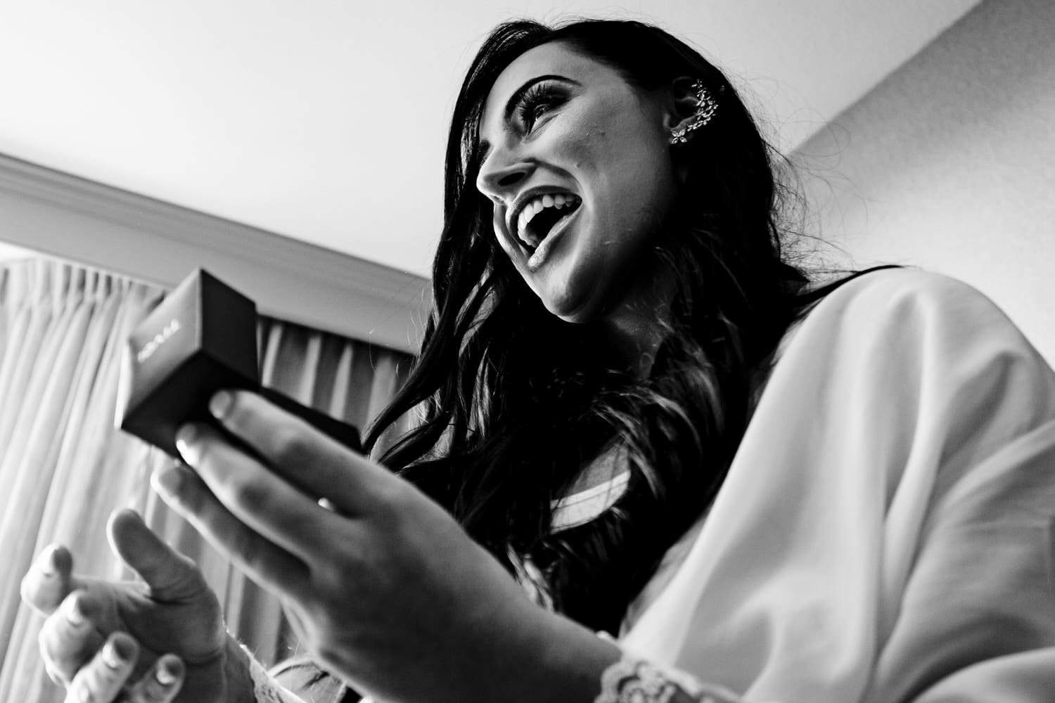 A candid black and white picture of a bride smiling excitedly in surprise as she opens diamond earrings from her groom. 