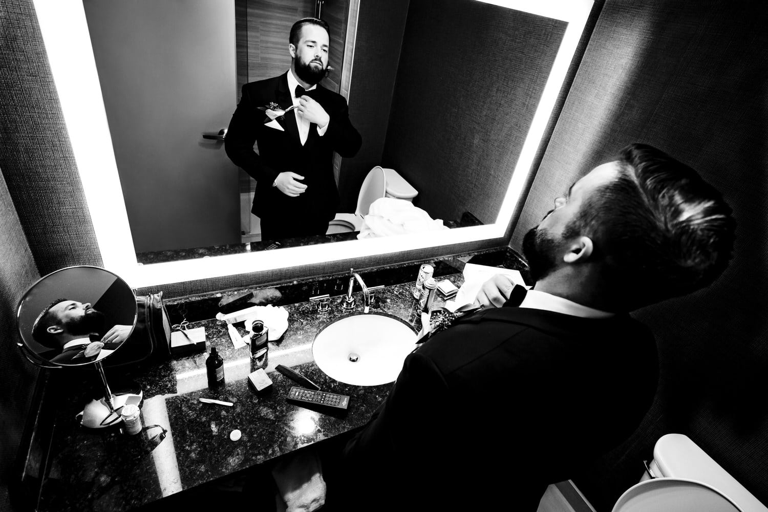 A candid black and white picture of a groom adjusting his bow tie in a bathroom mirror. 