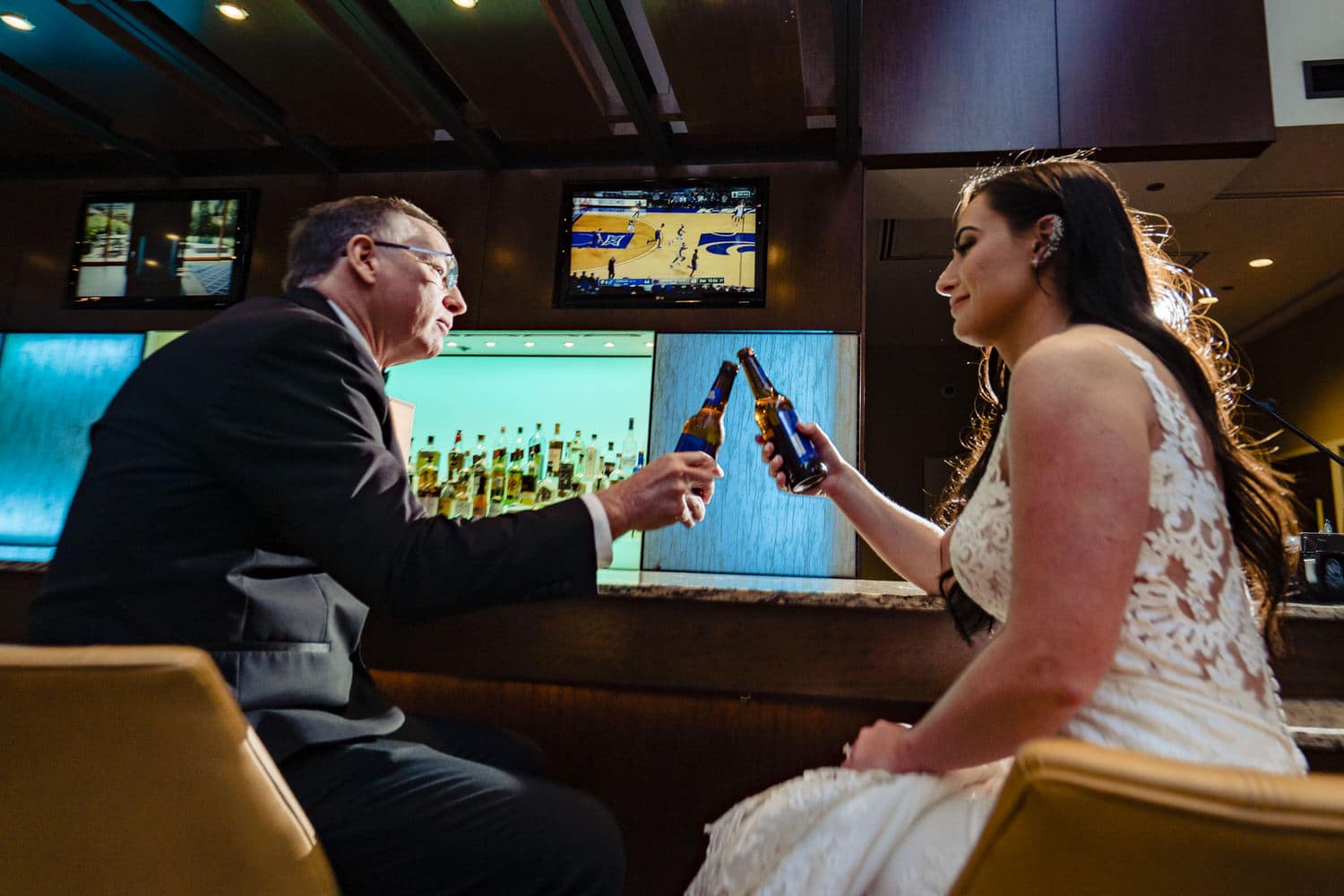 A colorful picture of a bride and her dad toasting with their beer bottles as they watch a KU game on a winter wedding day in Kansas City. 
