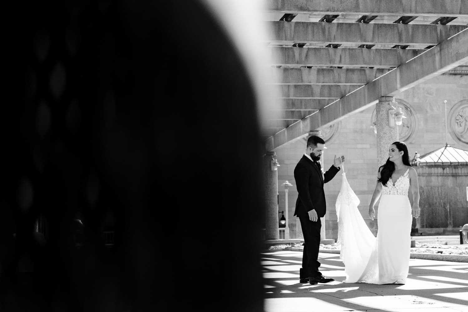 A candid black and white picture of a groom picking up the train of his bride's wedding gown. 