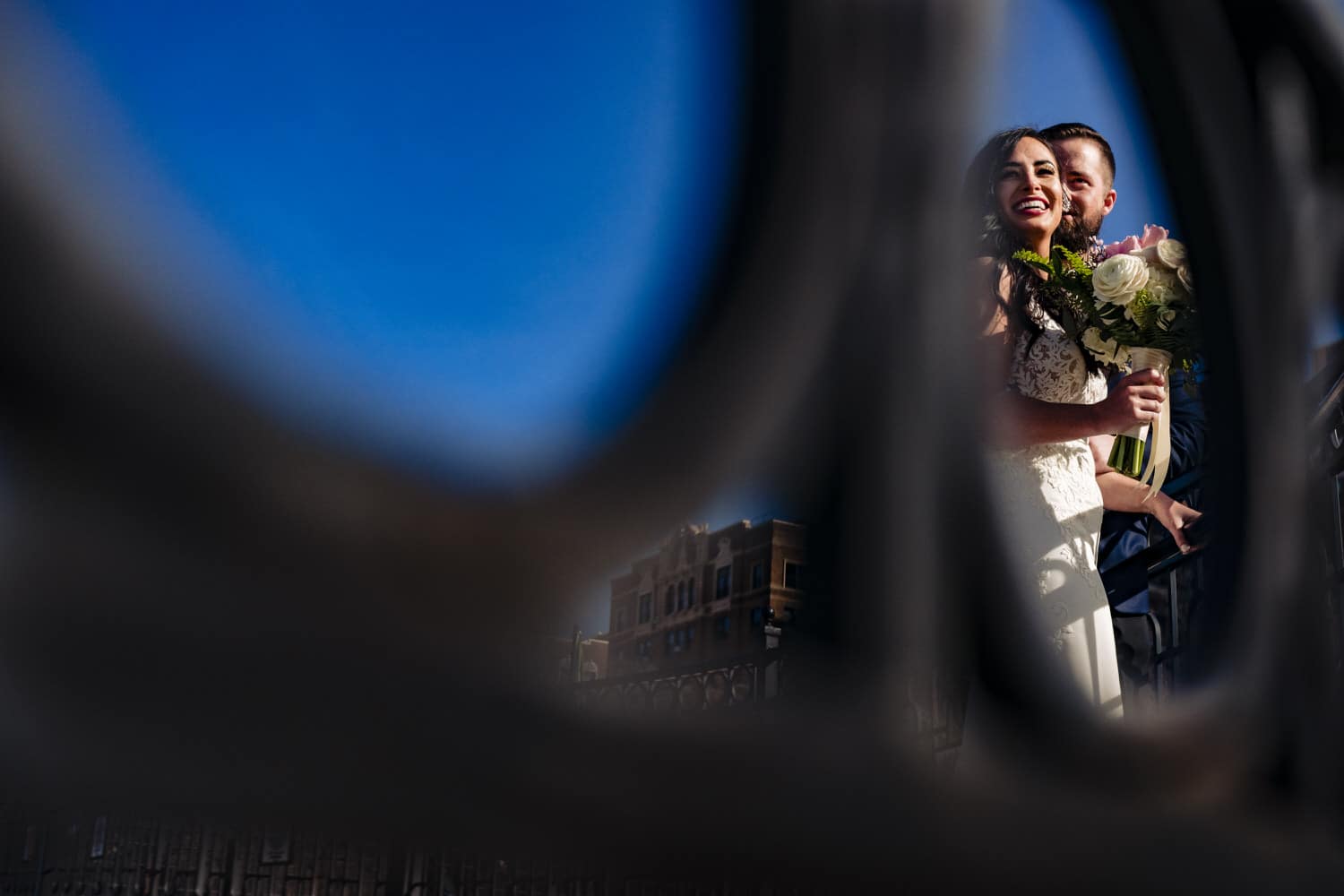 A colorful, candid picture taken through a fence line of a bride and group laughing and smiling together on a sunny winter day. 