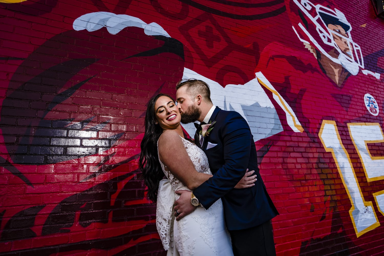 A colorful, candid portrait of a groom kissing his bride as they stand in front of a colorful Kansas City Chief's mural on their winter wedding day. 