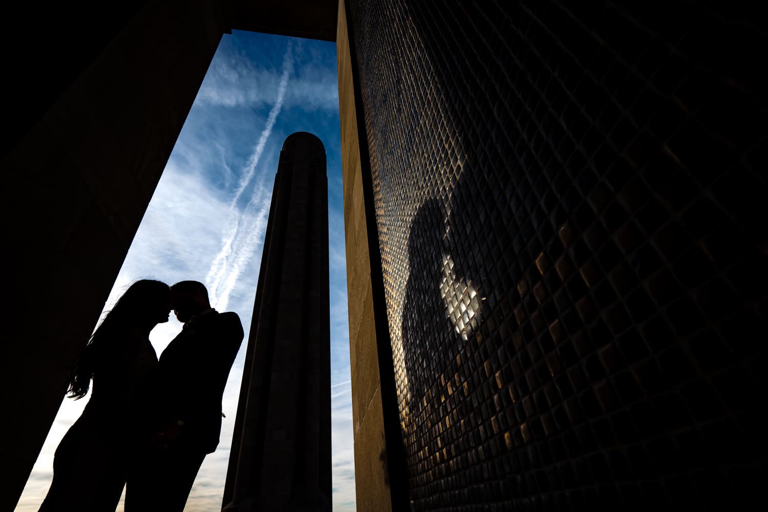 A silhouette of a bride and groom leaning in to share a kiss with the silhouette of Liberty Memorial visible in the background. 