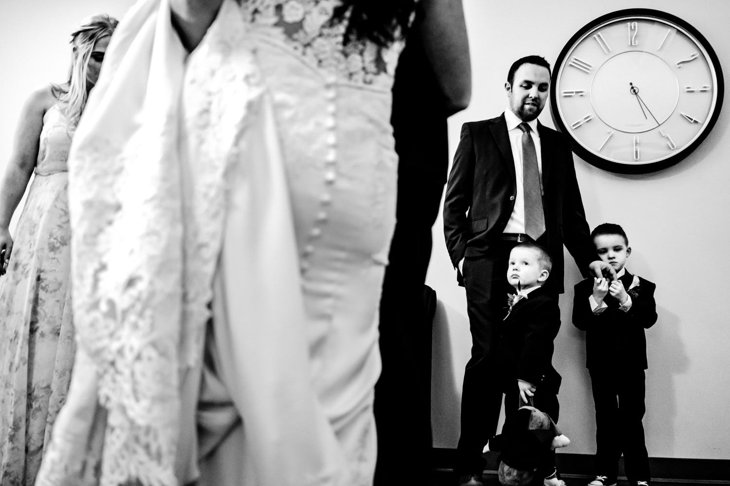 A candid black and white picture of two young ring bearers, standing underneath a giant clock, looking up at a bride. 