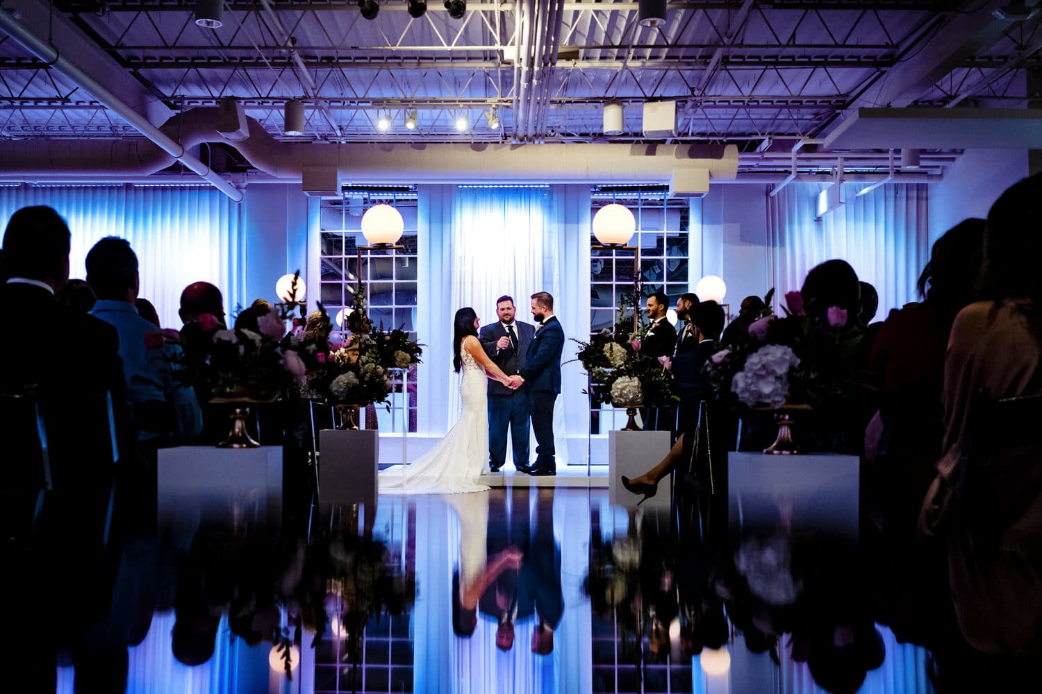 A colorful picture of a bride and groom holding hands and exchanging vows during a wedding ceremony at Pennway Place, their reflections visible on the floor in front of them on a winter day in Kansas City. 