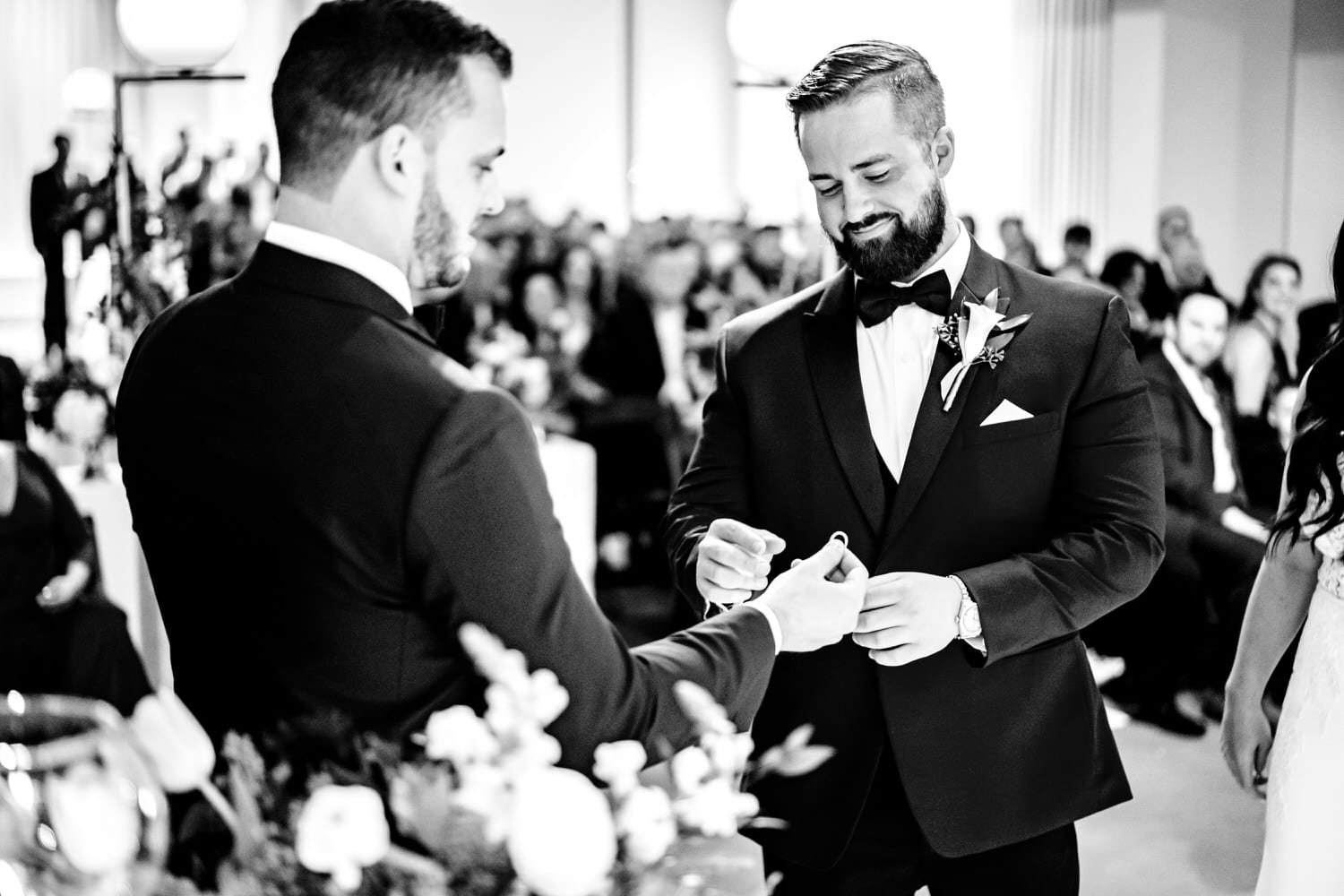A candid black and white picture of a groom smiling as he takes wedding bands from his best man during a wedding ceremony at Pennway Place in Kansas City. 