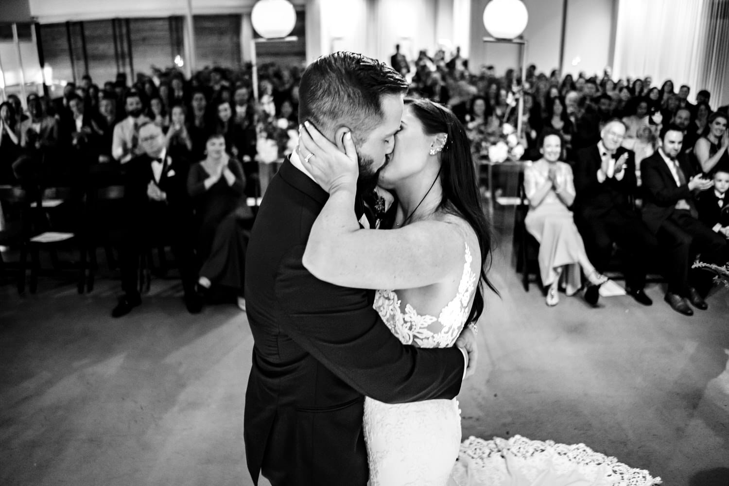 A candid black and white picture taken from behind the bride and groom, looking back at the audience as the bride and groom share their first kiss at the end of their wedding ceremony at Pennway Place in Kansas City. 