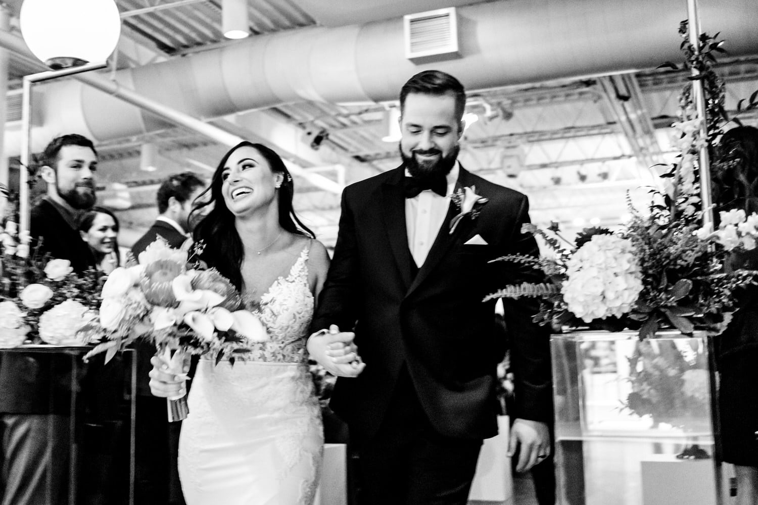 A candid black and white picture of a bride and groom smiling, laughing, and holding hands after their wedding ceremony at Pennway Place in Kansas City. 