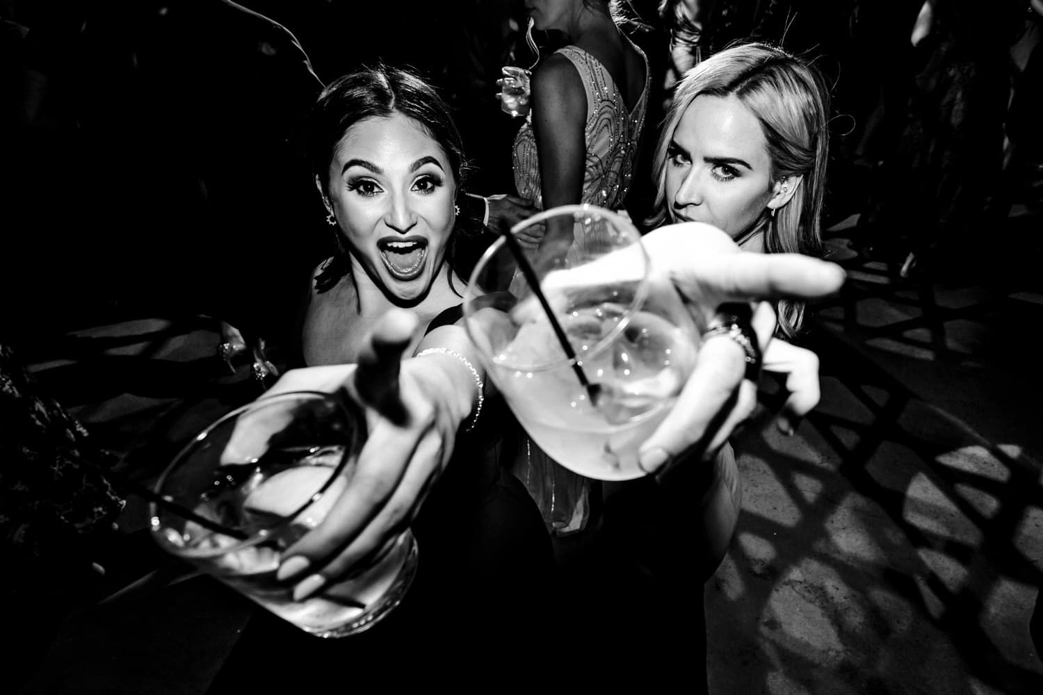 A candid black and white picture of two bridesmaids pointing their drink cups at the camera as they smile and laugh behind outstretched arms during a wedding reception at Pennway Place in Kansas City. 