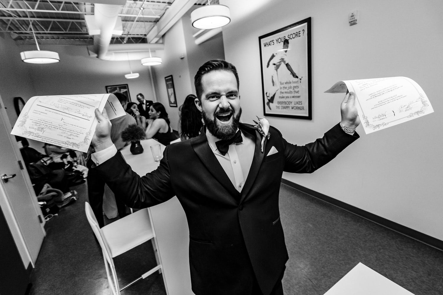 A candid black and white picture of a groom excitedly holding up two copies of a signed marriage license. 