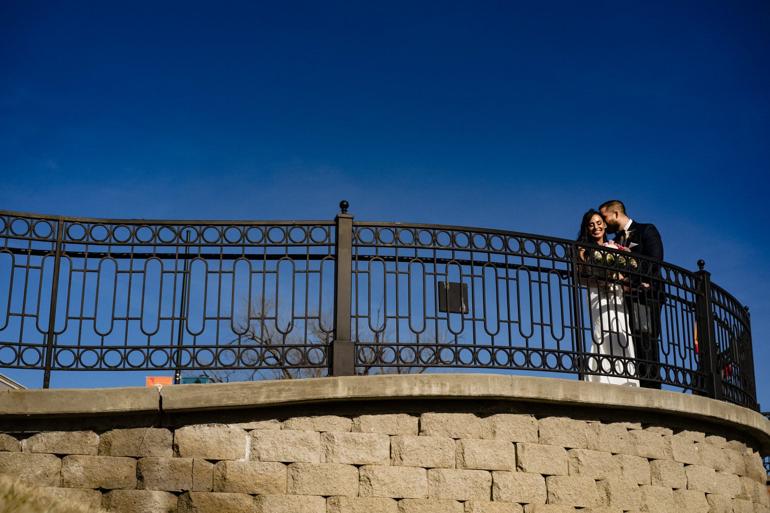 A colorful, wide portrait of a bride and groom standing behind a fence line on a sunny winter wedding day. 