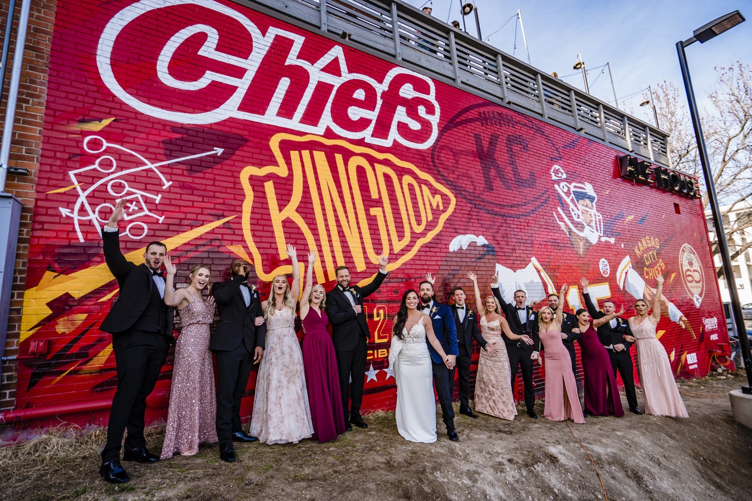A colorful, candid picture of a bride, groom, and their wedding party clapping and cheering as they stand in front of a Kansas City Chief's mural on the side of the Westport Alehouse on a winter wedding day. 
