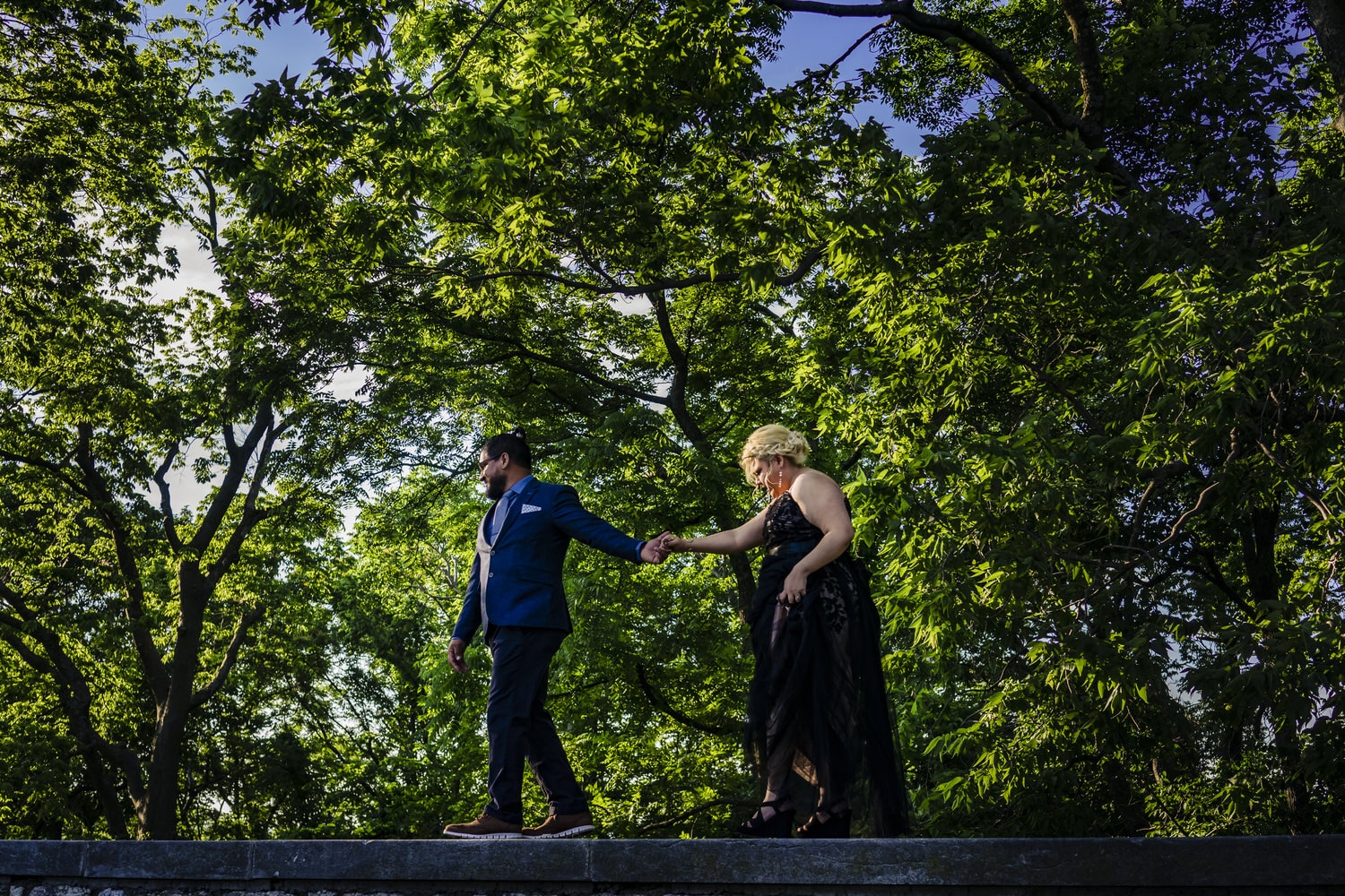 A colorful, candid picture of a man in a blue suit jacket leading a woman in a formal black dress across a path during their romantic engagement session in Kansas City. 
