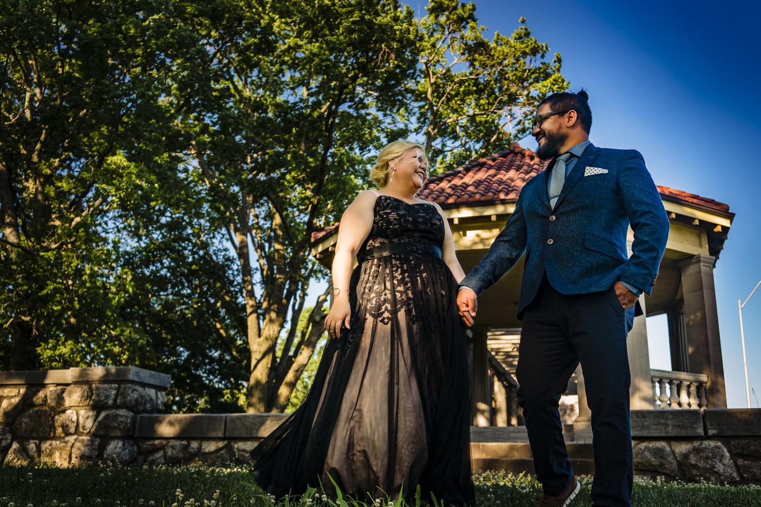 A colorful, candid picture of a man in a blue suit jacket leading a woman in a formal black dress across a path during their romantic engagement session in Kansas City. 