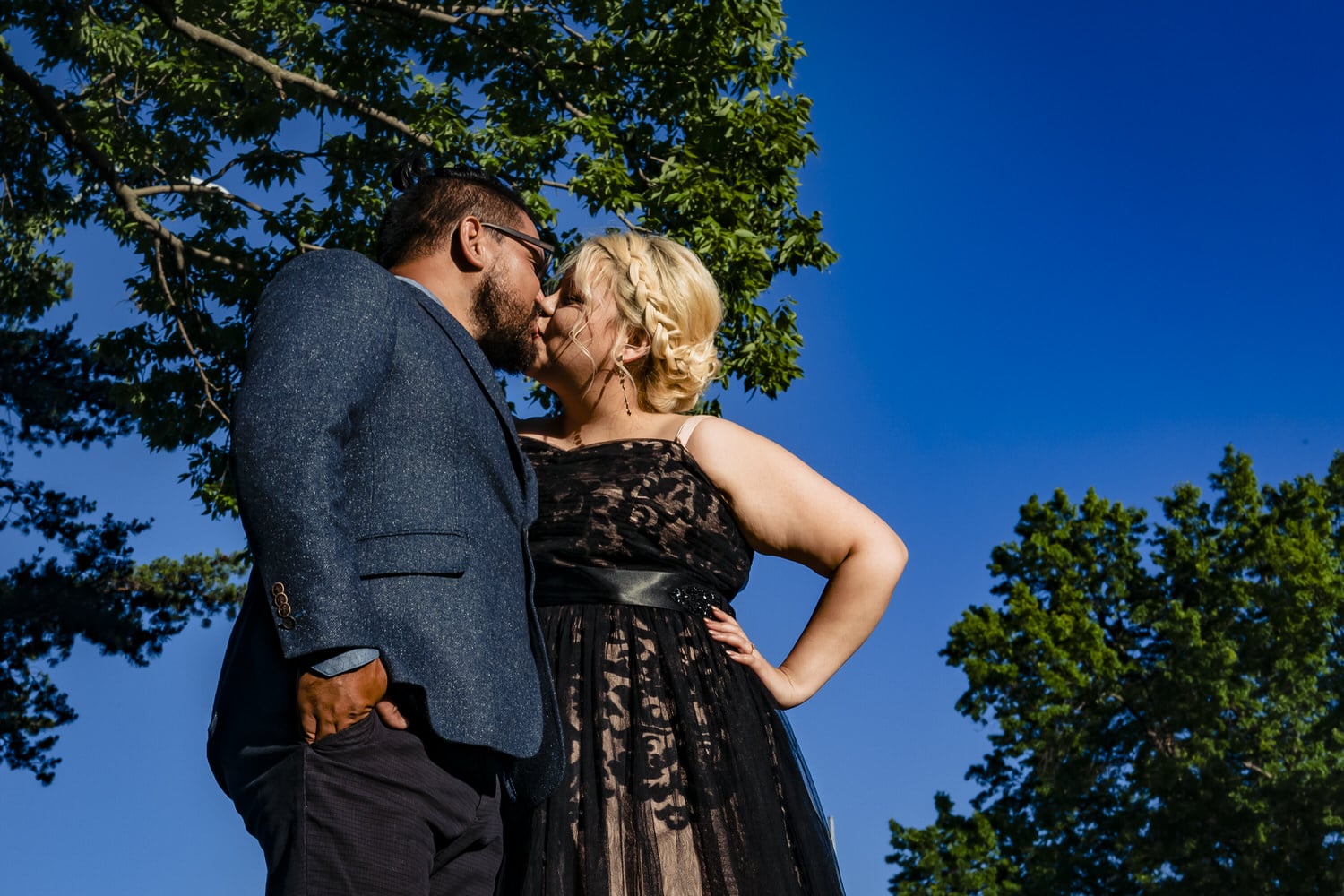 A candid picture of an engaged couple in formal wear leaning in to share a kiss against a bright blue backdrop of summer sky during an engagement session in Kansas City. 