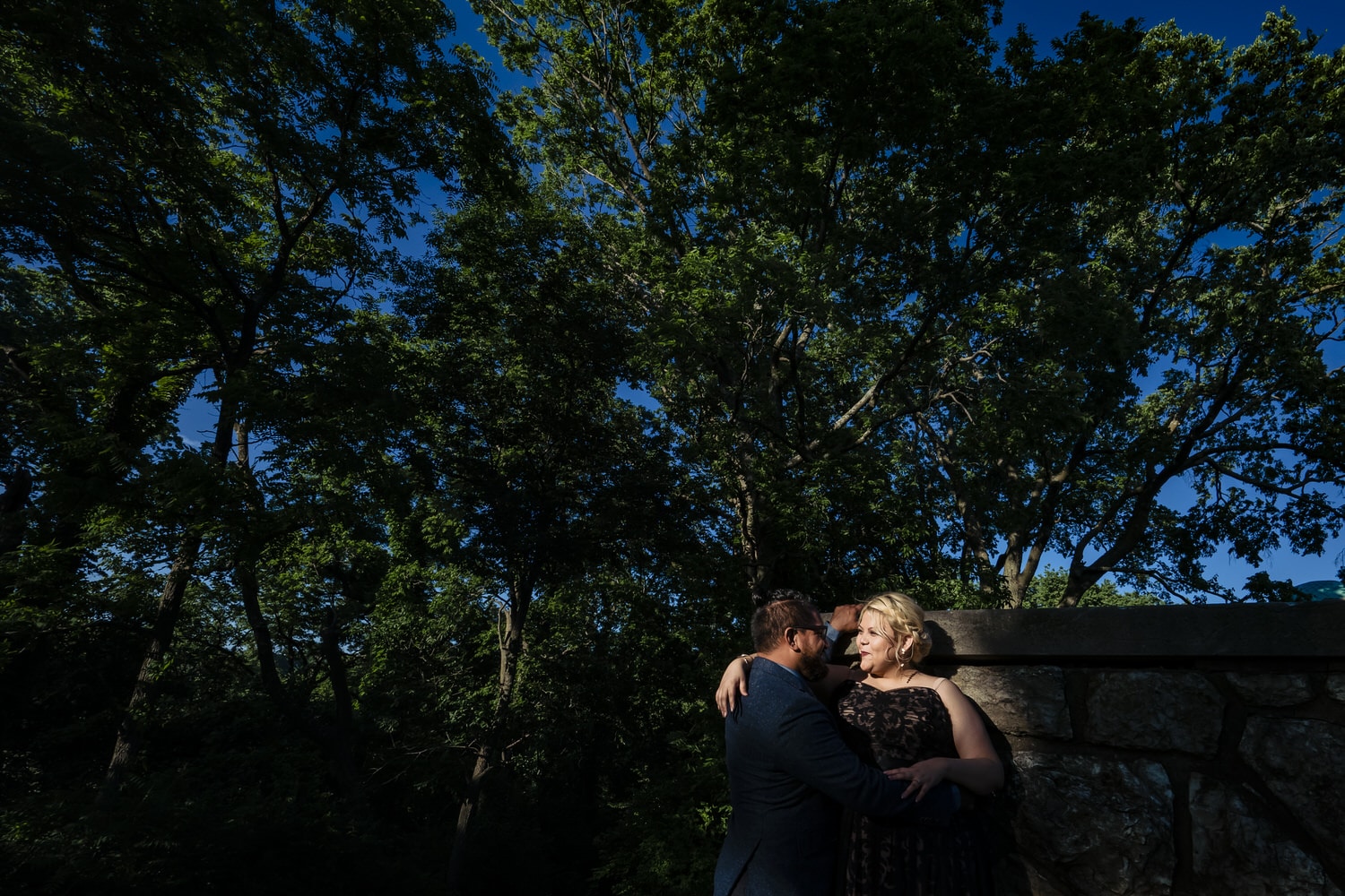 A wide, colorful picture of a man and woman leaning up against a stone wall, sharing an embrace during a romantic engagement session in Kansas City. 
