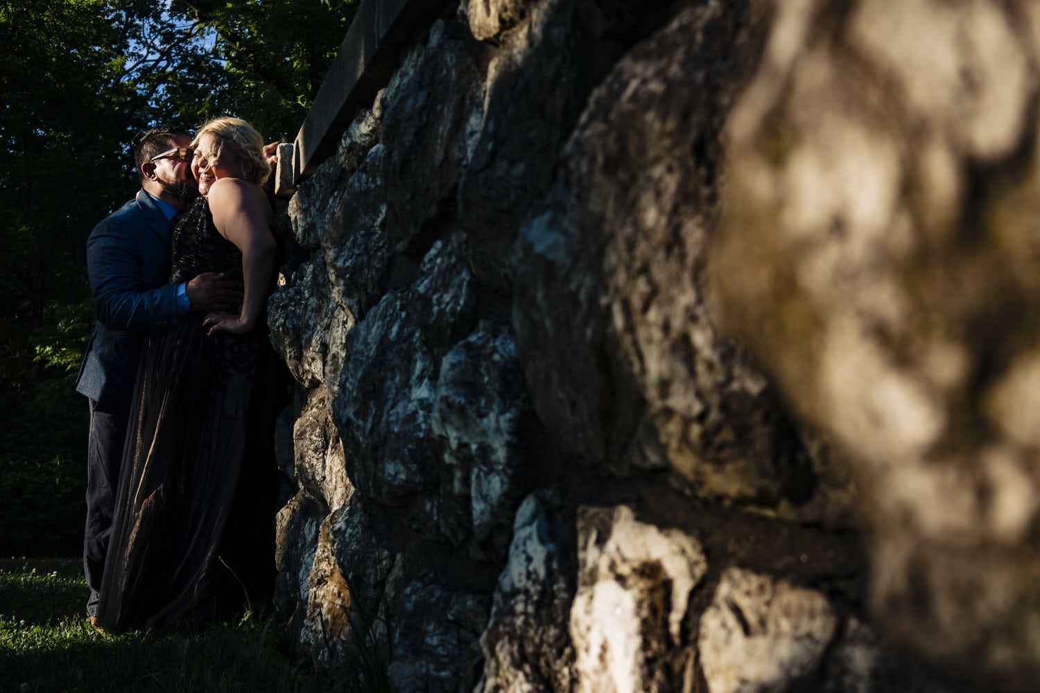 A wide, colorful picture of a man and woman leaning up against a stone wall, sharing an embrace during a romantic engagement session in Kansas City. 